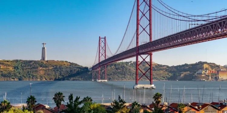 Il ponte 25 aprile di Lisbona con lo sfondo del Cristo Rei e il fiume Tejo, sono il confine di applicabilità di Uber Commute. Si vede un cielo azzurro e alcune piante.