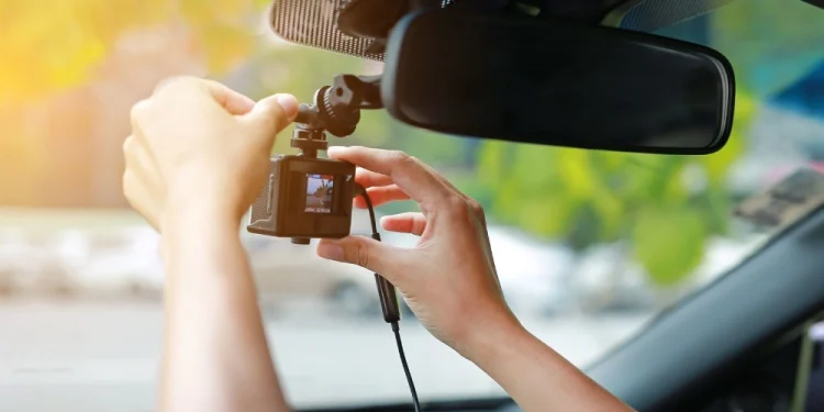 Una persona installa una dash cam sul parabrezza di un'auto in pieno sole, mostrando l'uso della dash cam in Portogallo, con vegetazione sfocata visibile all'esterno attraverso il finestrino.