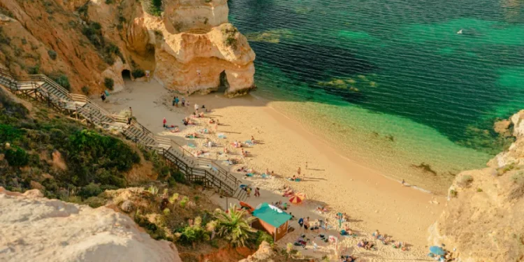 Veduta aerea di una Spiaggia sabbiosa in Algarve con acqua turchese, scogliere rocciose e scale di legno; le persone si godono una cara vacanza, prendendo il sole, nuotando e rilassandosi sotto gli ombrelloni vicino a una piccola struttura.