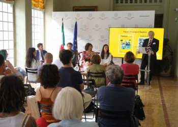 Un gruppo assiste a una presentazione di Traiettorie in una stanza luminosa evidenziando i temi della Guida psicologica su espatrio. L'ambasciatore Claudio Miscia è in piedi accanto a uno schermo che mostra una diapositiva, mentre altri siedono al tavolo. Il pubblico è rivolto verso di loro, con bandiere italiane e dell'Unione Europea sullo sfondo, evidenziando i temi della Guida psicologica espatrio.