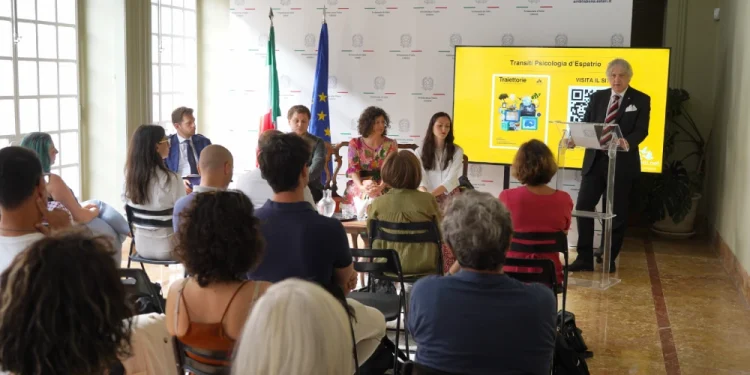 Un gruppo assiste a una presentazione di Traiettorie in una stanza luminosa evidenziando i temi della Guida psicologica su espatrio. L'ambasciatore Claudio Miscia è in piedi accanto a uno schermo che mostra una diapositiva, mentre altri siedono al tavolo. Il pubblico è rivolto verso di loro, con bandiere italiane e dell'Unione Europea sullo sfondo, evidenziando i temi della Guida psicologica espatrio.