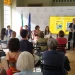 Un gruppo assiste a una presentazione di Traiettorie in una stanza luminosa evidenziando i temi della Guida psicologica su espatrio. L'ambasciatore Claudio Miscia è in piedi accanto a uno schermo che mostra una diapositiva, mentre altri siedono al tavolo. Il pubblico è rivolto verso di loro, con bandiere italiane e dell'Unione Europea sullo sfondo, evidenziando i temi della Guida psicologica espatrio.