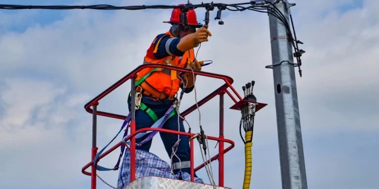 Un operaio con imbracatura di sicurezza e giubbotto arancione si trova su una piattaforma rialzata e sta riparando dei cavi elettrici su un palo della luce contro un cielo blu, forse per far fronte a un blackout 28 aprile.