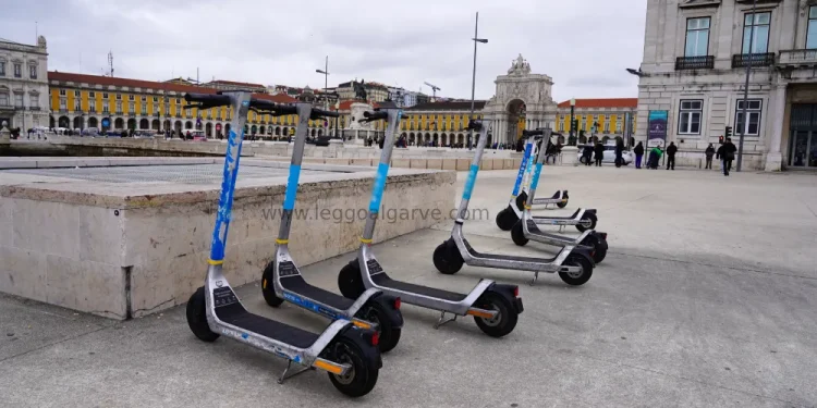 Quattro monopattini elettrici sono parcheggiati in fila su una piazza asfaltata, con edifici storici e persone sullo sfondo, probabilmente in attesa di controlli stradali sotto un cielo nuvoloso.