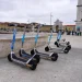 Quattro monopattini elettrici sono parcheggiati in fila su una piazza asfaltata, con edifici storici e persone sullo sfondo, probabilmente in attesa di controlli stradali sotto un cielo nuvoloso.