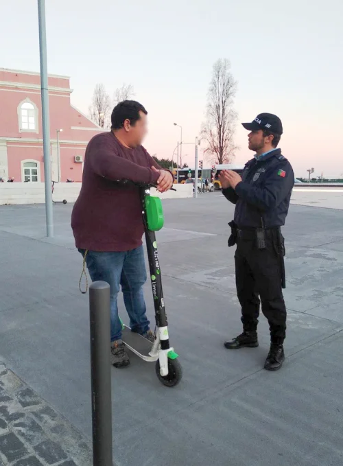 Un agente di polizia e un uomo discutono sulla strada durante uno dei controlli stradali su biciclette e monopattini a Lisbona. Si vedono sullo sfondo un palazzo rozzo e il cielo azzurro al tramonto.