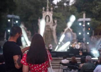 Un uomo e una donna si trovano tra il pubblico seduto a un concerto all'aperto durante la festa della città di Coimbra, con le luci luminose del palco, l'architettura in pietra ornata e gli alberi circostanti che esaltano la vivacità del luogo all'aperto.