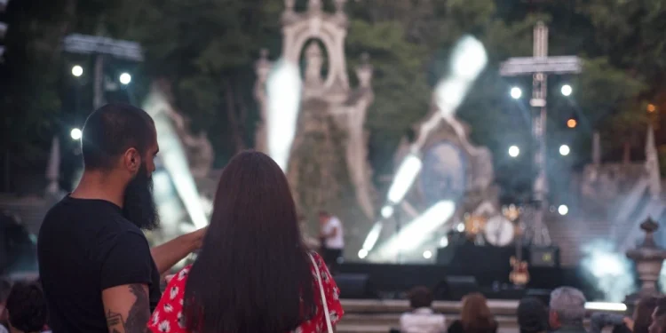Un uomo e una donna si trovano tra il pubblico seduto a un concerto all'aperto durante la festa della città di Coimbra, con le luci luminose del palco, l'architettura in pietra ornata e gli alberi circostanti che esaltano la vivacità del luogo all'aperto.