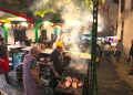 In una piazza guida durante le feste dei santi popolari di Lisbona due anziani grilgliano del cibo. Si vedono altre persone, degli addobbi e delle piante.