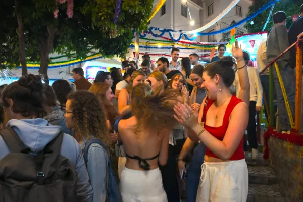 Molta gente in strada durante i festeggiamenti dei Santi Popolari a Lisbona. Si vedono bancarelle e addobbi colorati e in un angolo le foglie di una pianta.