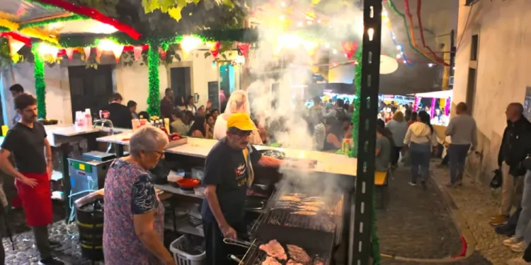 In una piazza guida durante le feste dei santi popolari di Lisbona due anziani grilgliano del cibo. Si vedono altre persone, degli addobbi e delle piante.