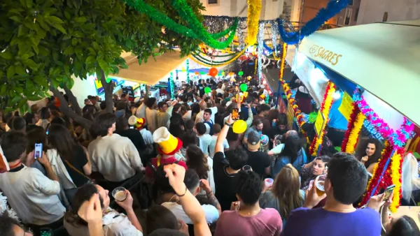 Molta gente in strada durante i festeggiamenti dei Santi Popolari a Lisbona. Si vedono bancarelle e addobbi colorati e in un angolo le foglie di una pianta. 