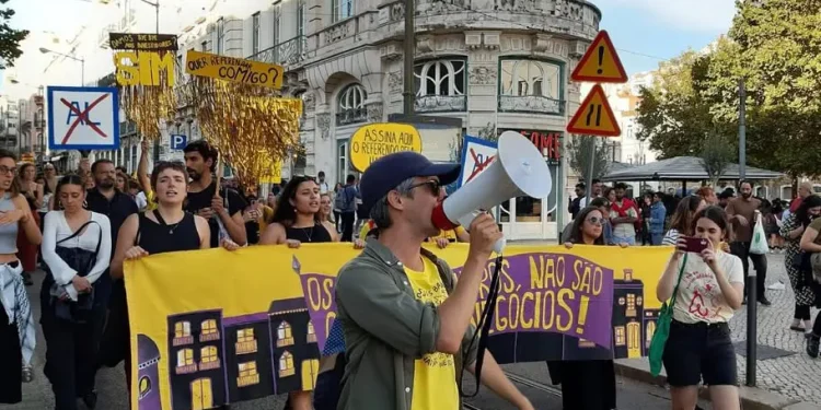Un gruppo di persone marcia in una strada della città durante le manifestazioni per il diritto alla casa, tenendo in mano striscioni e cartelli, alcuni dei quali recitano "SIM" e "ASSINA AQUI O REFERENDO!", mentre una persona in testa parla in un megafono. Alle loro spalle sono visibili edifici e alberi.