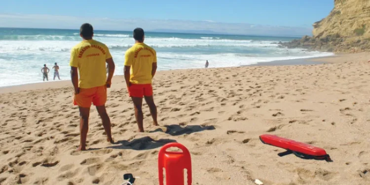Due bagnini in maglietta gialla e pantaloncini rossi sono in piedi su una spiaggia sabbiosa in Portogallo, a sorvegliare le persone in mare per prevenire morti per annegamento. L'attrezzatura di salvataggio è nelle vicinanze, con cielo terso e scogliere sulla destra.