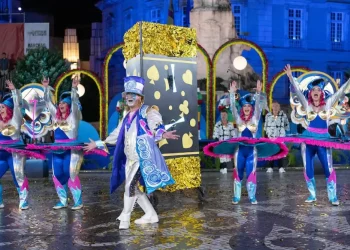 Artisti in costumi colorati ed elaborati, che raffigurano temi come carte da gioco e farfalle, danzano con le braccia alzate davanti a un grande oggetto di carte durante la vivace sfilata notturna di Sant'Antonio Lisbona.