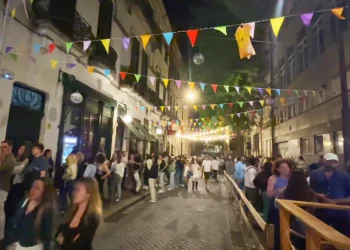 Una vivace scena di strada di notte a Porto con persone riunite sotto colorati festoni triangolari dove è vietata la vendita di alcolici. La strada di ciottoli è affollata e illuminata dai lampioni.
