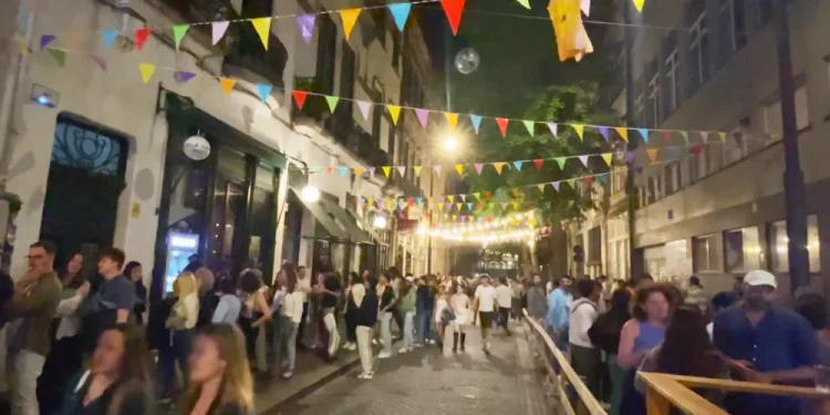 Una vivace scena di strada di notte a Porto con persone riunite sotto colorati festoni triangolari dove è vietata la vendita di alcolici. La strada di ciottoli è affollata e illuminata dai lampioni.