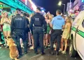 Tre agenti di polizia e un cane si trovano di fronte a una vivace folla di giovani all'esterno di un bar illuminato di notte, sorvegliando la movida nell'ambito del posto di controllo GNR di Albufeira tra luci al neon e vivaci scene da marciapiede.