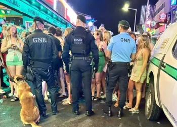 Tre agenti di polizia e un cane si trovano di fronte a una vivace folla di giovani all'esterno di un bar illuminato di notte, sorvegliando la movida nell'ambito del posto di controllo GNR di Albufeira tra luci al neon e vivaci scene da marciapiede.