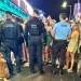 Tre agenti di polizia e un cane si trovano di fronte a una vivace folla di giovani all'esterno di un bar illuminato di notte, sorvegliando la movida nell'ambito del posto di controllo GNR di Albufeira tra luci al neon e vivaci scene da marciapiede.