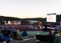 Persone sedute su sedie a sdraio e sacchi di fagioli all'aperto al tramonto, mentre guardano un musicista dal vivo sul palco del Lagoa Jazz Festival 2024. Le grandi lettere "LAGOA" e il fiume creano uno sfondo scenografico per l'evento.