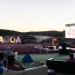 Persone sedute su sedie a sdraio e sacchi di fagioli all'aperto al tramonto, mentre guardano un musicista dal vivo sul palco del Lagoa Jazz Festival 2024. Le grandi lettere "LAGOA" e il fiume creano uno sfondo scenografico per l'evento.