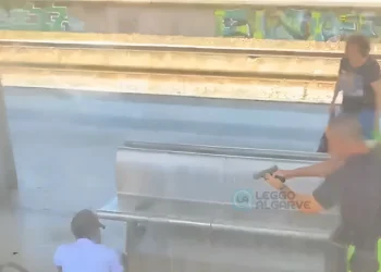 Un poliziotto punta una pistola contro un individuo reo di minacce su un treno vicino a una panchina di metallo alla stazione di Lisbona, mentre un terzo passeggero si trova nelle vicinanze. Su un muro sono visibili dei graffiti e sullo sfondo i binari del treno.