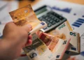 Alcune banconote di euro e una calcolatrice con delle bollette per valutare se è possibile vivere con il salario minimo in Portogallo. Una mano mantiene una banconota da 50 euro.