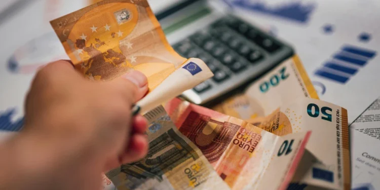 Alcune banconote di euro e una calcolatrice con delle bollette per valutare se è possibile vivere con il salario minimo in Portogallo. Una mano mantiene una banconota da 50 euro.