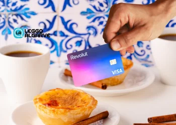 Una mano tiene una carta di credito con logo revolut sullo sfondo di prodotti simbolo del Portogallo, come caffè e pastel de nata e azulejo.