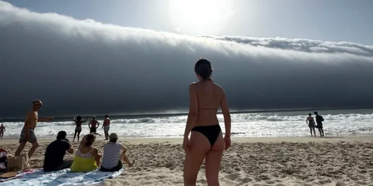 Un folto gruppo di persone osserva delle spettacolari nuvole su una spiaggia in Portogallo. In primo piano una donna in costume di spalle.