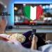Una donna in Portogallo mentre guardare una tv con bandiera italiana in salotto con un telecomando e dei pop-corn in mano