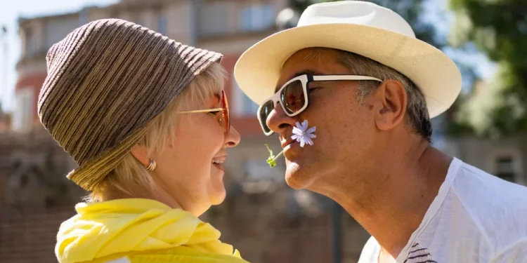 Due pensionati italiani sono faccia a faccia e sembrano felici di essere all estero, hanno cappelli in testa, occhiali e l'uomo un fiore viola in bocca. Sullo sfondo sfuocata una casa e delle piante.