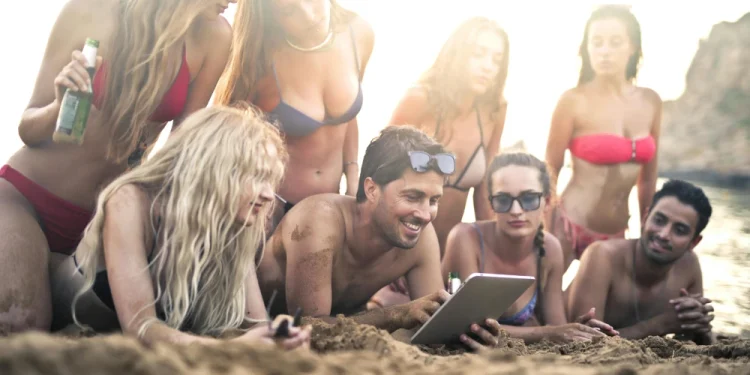 Un gruppo di ragazzi su una spiaggia in Portogallo mentre utilizzano un tablet per leggere le regole in vigore. Sono tutti in costume e sono sorridenti c'è una ragazza con una birra in mano, si vede sullo sfondo il mare e gli scogli.