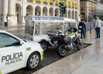 Un auto della polizia effettua dei controlli su tuk-tuk per verificare se irregolari su una strada di Lisbona. Si vede un palazzo con delle colonne sullo sfondo.
