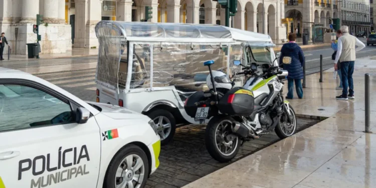 Un auto della polizia effettua dei controlli su tuk-tuk per verificare se irregolari su una strada di Lisbona. Si vede un palazzo con delle colonne sullo sfondo.