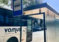 Un autobus Vamus bianco è fermo a una fermata riparata sotto un cartello blu in una giornata di sole, con auto parcheggiate ed edifici visibili sullo sfondo: l'ideale per i viaggiatori che utilizzano l'Abbonamento Passe Algarve.
