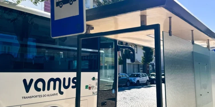 Un autobus Vamus bianco è fermo a una fermata riparata sotto un cartello blu in una giornata di sole, con auto parcheggiate ed edifici visibili sullo sfondo: l'ideale per i viaggiatori che utilizzano l'Abbonamento Passe Algarve.
