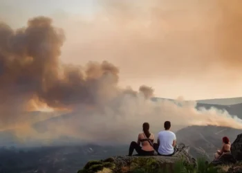 Tre persone sono sedute di spalle osservando alcuni incendi nelle foreste in Portogallo, discutendo sulle probabili cause. Sullo sfondo si vede una fitta coltre di fumo invadere delle montagne.