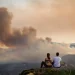 Tre persone sono sedute di spalle osservando alcuni incendi nelle foreste in Portogallo, discutendo sulle probabili cause. Sullo sfondo si vede una fitta coltre di fumo invadere delle montagne.