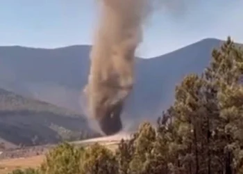 Un grande Diavolo di Sabbia a Covilhã, o diavolo di polvere, si alza dal suolo, facendo vorticare polvere e detriti in aria sullo sfondo di colline e alberi sotto un cielo limpido.