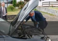 Il Presidente della Repubblica portoghese Marcelo osserva in modo pragmatico dentro al motore in avaria della sua auto, con un uomo della scorta che osserva la strada. Sullo sfondo si vede un giardino ed una costruzione.