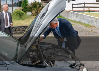 Il Presidente della Repubblica portoghese Marcelo osserva in modo pragmatico dentro al motore in avaria della sua auto, con un uomo della scorta che osserva la strada. Sullo sfondo si vede un giardino ed una costruzione.