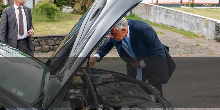 Il Presidente della Repubblica portoghese Marcelo osserva in modo pragmatico dentro al motore in avaria della sua auto, con un uomo della scorta che osserva la strada. Sullo sfondo si vede un giardino ed una costruzione.
