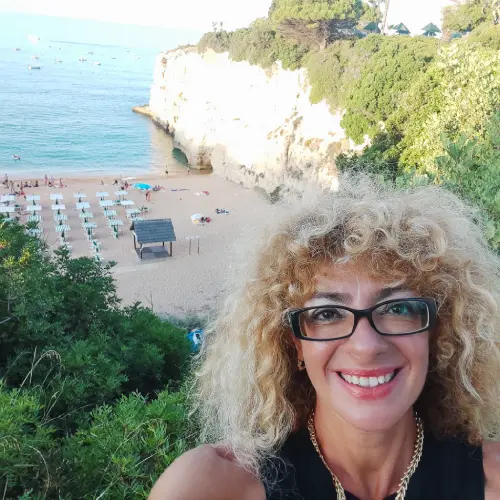 Silvana Di Liberto, sorridente posa di fronte ad una spiaggia in Algarve con alcune sdraio e della vegetazione. 