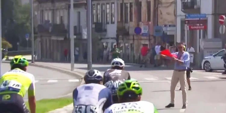 Alcuni ciclisti della Volta a Portugal vengono fermati da un uomo con bandiera rossa nel mezzo di una strada cittadina.
