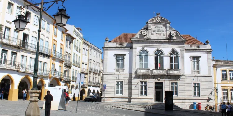 Nella piazza di Evora il palazzo del Banco de Portugal circondato da edifici portoghesi storici con altre banche, una strada non trafficata e alcune persone. Il cielo è sgombro dalle nuvole.