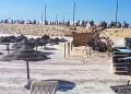Alcune onde del mare devastano la spiaggia di Caparica, si vedono ombrelloni e sdraio da spiaggia immerse nell'acqua. Sullo sfondo sopra una duna di sabbia alcune persone .