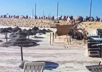 Alcune onde del mare devastano la spiaggia di Caparica, si vedono ombrelloni e sdraio da spiaggia immerse nell'acqua. Sullo sfondo sopra una duna di sabbia alcune persone .
