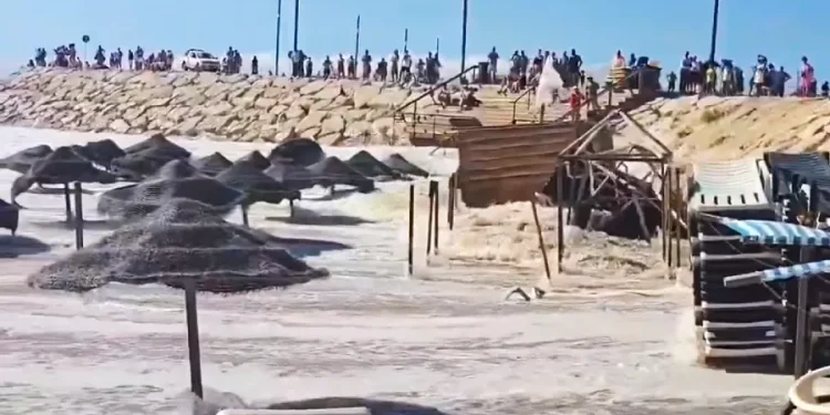 Alcune onde del mare devastano la spiaggia di Caparica, si vedono ombrelloni e sdraio da spiaggia immerse nell'acqua. Sullo sfondo sopra una duna di sabbia alcune persone .